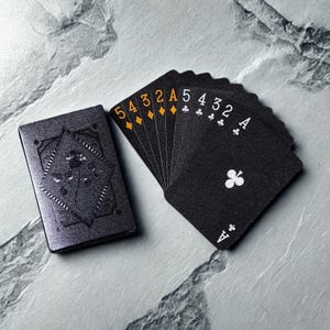 May include: A deck of playing cards with black card faces and white and gold markings. The cards are fanned out next to a matching black card case with an embossed floral design. The suits and numbers are visible on the cards.