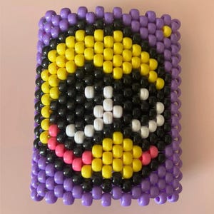 Puede incluir: Una creación rectangular de cuentas con un diseño de carita sonriente. La cara está hecha con cuentas amarillas, negras, blancas y rosas, sobre un borde morado. Las cuentas están dispuestas para crear una superficie texturizada.