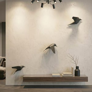 Pode incluir: Um design de interiores moderno com três esculturas geométricas de pássaros montadas em uma parede texturizada. Uma prateleira flutuante de madeira escura com livros, um vaso e um galho decorativo. Uma luminária preta no teto.