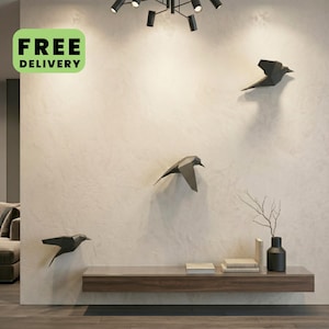 Könnte beinhalten: Drei schwarze geometrische Vogel-Wandskulpturen an einer strukturierten, cremeweißen Wand. Die Vögel sind in einem versetzten Flugmuster angeordnet. Darunter befindet sich ein langes, horizontales Holzregal mit Büchern und einer schwarzen Vase mit Zweigen.