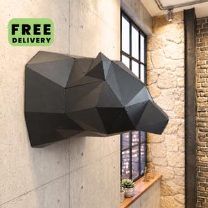 Könnte beinhalten: Eine Wandskulptur aus mattschwarzem Metall in Form eines geometrischen Bärenkopfes. Das Objekt besteht aus mehreren flachen, kantigen Paneelen, die mit sichtbaren Nieten verbunden sind und eine facettierte Optik an einer Betonwand bilden.