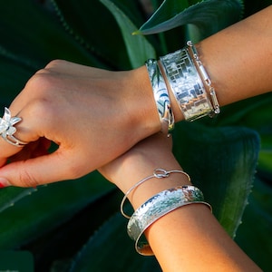 Peut inclure: Gros plan sur des mains ornées de bijoux en argent. Une bague fleur et plusieurs bracelets sont visibles, dont un jonc martelé, un bracelet mosaïque et un bracelet en forme de nœud. Le fond est composé de feuillage vert.