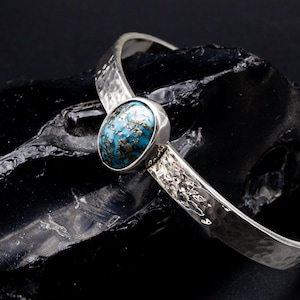Peut inclure: Un bracelet manchette en argent avec une texture martelée et une pierre centrale de forme ovale présentant un motif tacheté bleu et noir. La bande métallique possède une finition polie et une large ouverture, mettant en valeur le contraste entre l'argent brillant et la pierre sombre.