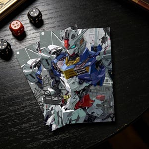 Op de afbeelding: Twee trading card sleeves met een gedetailleerde illustratie van een Gundam robot in blauw, wit, rood en geel. De kaarten liggen op een donkere ondergrond met dobbelstenen en een boek op de achtergrond. De robot staat tegen een stadsgezicht.