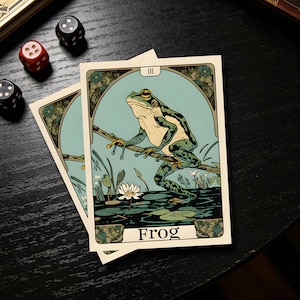 May include: Two tarot cards featuring a green frog perched on a branch, set against a light blue background. The word "Frog" is printed at the bottom. The cards have an art nouveau style with floral accents and are placed on a dark surface.