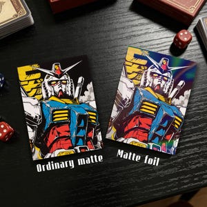 May include: Two trading cards featuring a colorful illustration of a robot. One card is labeled "Ordinary matte" and the other "Matte foil." The robot is primarily blue, red, yellow, and white, with a detailed design. The cards are on a black surface.