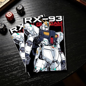 Puede incluir: Dos pegatinas con una ilustraci&oacute;n detallada de un robot blanco, negro y verde azulado con el texto "RX-93" y "Gundam" sobre un fondo negro. Las pegatinas est&aacute;n sobre una superficie oscura con dados.