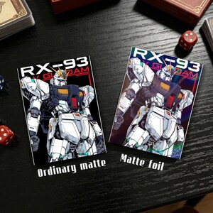 Puede incluir: Dos tarjetas rectangulares con una ilustraci&oacute;n de robot y el texto "RX-93 GUNDAM". Una tarjeta dice "Ordinary matte" y la otra "Matte foil". El robot es blanco, negro y rojo. Dados y una caja de cartas en el fondo.