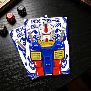May include: Two cards featuring a detailed illustration of a Gundam robot, primarily in blue, red, white, and yellow. The cards have the text "RX 78-2" and "EFSF". Two dice, one red and one black, and another black die are visible.