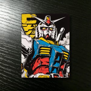 May include: A colorful illustration of a Gundam robot. The robot is depicted in a dynamic pose with a detailed design, featuring red, blue, yellow, and white accents against a black background. The image has a bold, graphic style.
