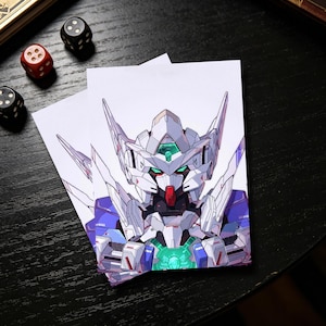 Op de afbeelding: Twee witte kaarten met een gedetailleerde illustratie van een Gundam-robot. De robot is overwegend wit met blauwe en rode accenten en een groene kern. Dobbelstenen liggen verspreid rond de kaarten op een zwart oppervlak.