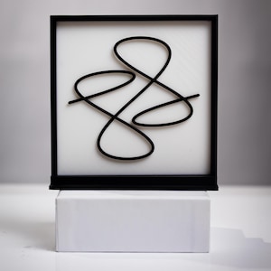 May include: A square black frame containing a white background with a raised, black, continuous looping line design centered within the frame. The frame is positioned on a solid white rectangular base, creating a clean, high-contrast display.