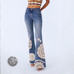 Könnte beinhalten: Blaue Jeans aus Denim mit hoher Taille und ausgestelltem Beinverlauf. Der untere Teil der Beine ist mit großen, kreisförmigen, cremefarbenen Häkelspitzen-Applikationen in Form von Deckchen und kleinen floralen Häkelakzenten verziert.