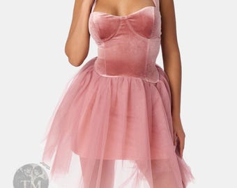Può includere: Un abito corto in velluto rosa polvere con un corpetto strutturato stile corsetto, scollo a cuore e spalline larghe. La gonna è composta da diversi strati di tulle trasparente e voluminoso che crea una silhouette ampia e svasata.
