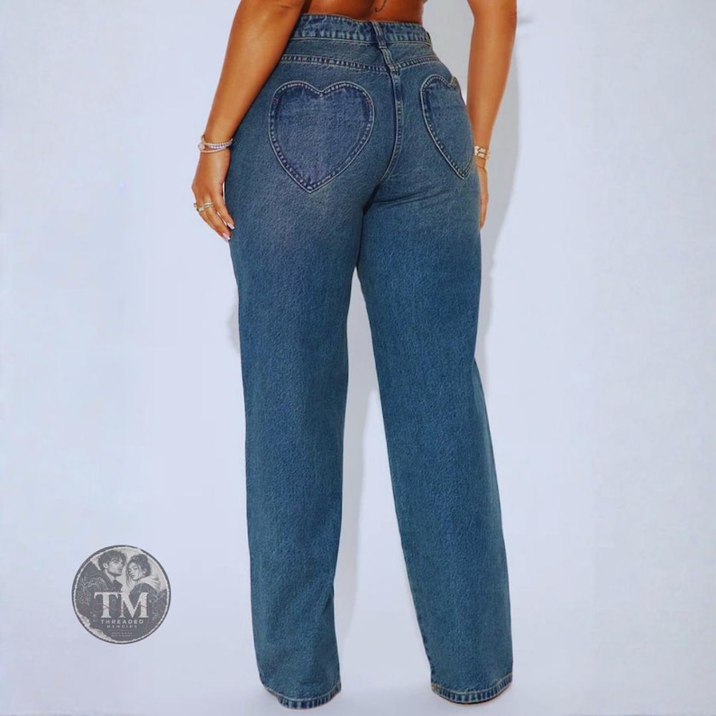 May include: Medium-wash blue denim jeans featuring a high-waisted fit and two prominent heart-shaped back pockets. The jeans have a straight-leg silhouette and a classic button and zipper fly closure. A circular logo with the text TM Threaded is visible.