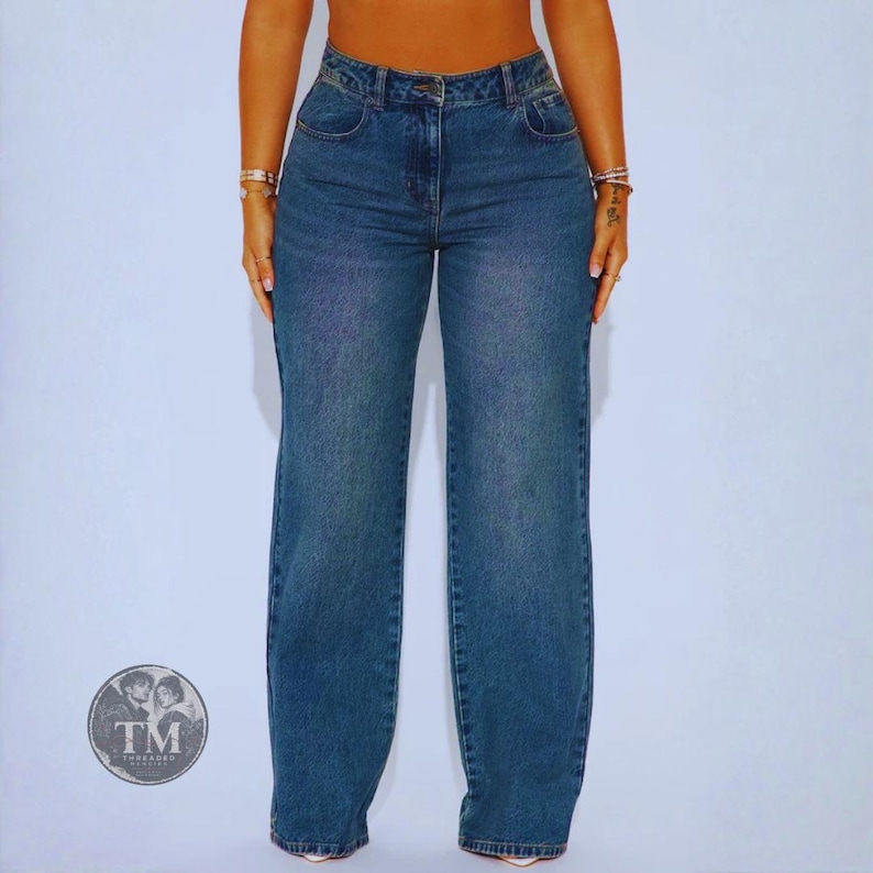 May include: High-waisted blue denim jeans featuring a wide-leg silhouette, a classic button and zipper fly closure, and traditional five-pocket styling. The medium-wash fabric displays subtle fading along the thighs and seams for a textured look.