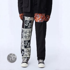 Op de afbeelding: Zwarte broek met een contrasterend ontwerp. De linkerpijp heeft een wit en zwart bloemrijk bandana-stijl patroon in vierkante panelen. De rechterpijp is effen zwart met een oranje en blauwe bloemrijke opgestikte zak op de heup.