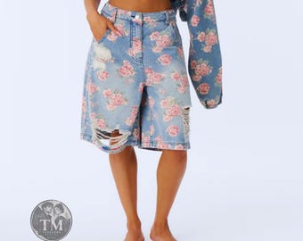 Può includere: Pantaloncini in denim azzurro con motivo floreale di rose rosa, vita alta, chiusura con bottoni e zip e dettagli usurati sulla coscia destra. Una manica di giacca in denim abbinata con lo stesso motivo floreale è tenuta di lato.