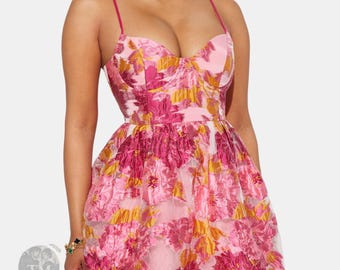 Può includere: Un abito senza maniche con scollo a cuore e spalline sottili. Il tessuto presenta una base rosa chiaro con un vivace motivo floreale nelle tonalità magenta, rosa scuro e oro, creando un aspetto strutturato in stile jacquard.