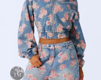 Può includere: Un completo composto da una giacca in denim azzurro e pantaloni a vita alta abbinati, caratterizzati da un motivo floreale con rose rosa. La giacca presenta una chiusura a zip frontale, colletto e maniche lunghe. I pantaloni hanno chiusura a zip.