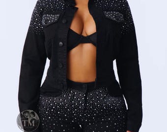 Può includere: Una giacca di jeans nera con colletto, maniche lunghe e chiusura frontale con bottoni. La giacca è decorata con strass color argento concentrati sulle spalle e sulle tasche sul petto. Un reggiseno nero è visibile sotto la giacca aperta.