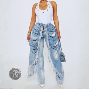 May include: A white sleeveless bodysuit with silver grommet details around the neckline paired with light-wash blue denim jeans featuring dramatic vertical ruffles, frayed edges, and a distressed finish. A small blue handbag is held at the side.