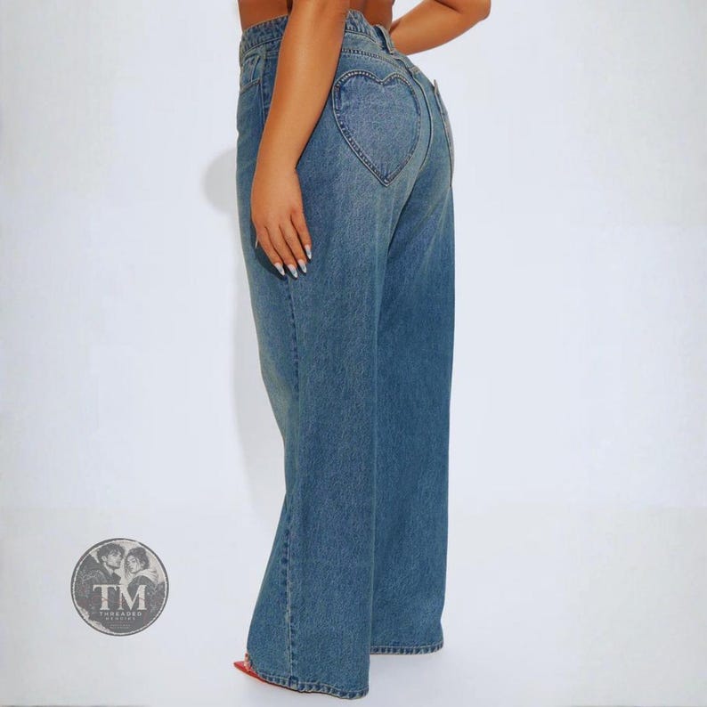 May include: Blue denim jeans featuring a high-waisted fit and wide-leg silhouette. The back of the jeans includes a prominent heart-shaped pocket design stitched onto the rear. The denim has a medium wash finish with visible seams and belt loops.