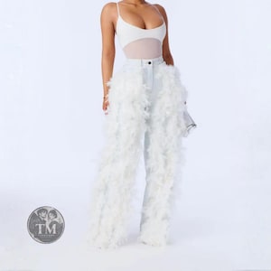 May include: A white bodysuit with thin straps paired with light blue denim jeans featuring long, voluminous white feather trim along the legs. The jeans have a classic button and zipper fly closure. A circular logo with the text TM Threaded is visible.