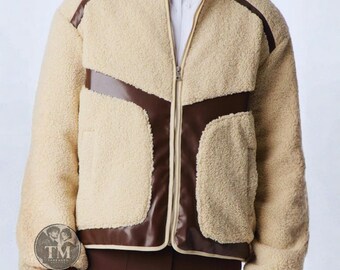 Può includere: Una giacca in pile sherpa color crema con inserti in similpelle marrone scuro sul petto e sulle spalle. La giacca presenta una chiusura a zip frontale, un colletto rialzato e maniche lunghe. È visibile un logo circolare con il testo TM.