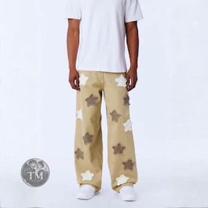 Può includere: Pantaloni a gamba larga color beige con molteplici toppe a forma di stella dai bordi sfrangiati in bianco e marrone scuro. I pantaloni hanno una vestibilità rilassata e sono abbinati a una t-shirt bianca semplice e scarpe da ginnastica bianche. È visibile un logo circolare con il testo TM Threaded.