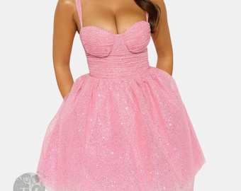 Può includere: Un miniabito rosa glitterato caratterizzato da uno scollo a cuore con corpetto arricciato e spalline sottili. La gonna è voluminosa e svasata, realizzata con strati di tessuto in tulle scintillante che catturano la luce.