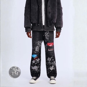 May include: Black denim pants featuring a distressed grey paint splatter pattern, a red patch with a car graphic, and a rectangular patch with the text ART THROUGH ANARCHY. The pants are paired with a dark grey denim jacket and white sneakers.
