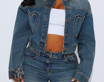 May include: A cropped blue denim jacket featuring a collar, long sleeves, and a button-down front. The jacket is detailed with prominent heart-shaped stitching on the chest panels. It is worn over a plain white cropped top with matching blue denim jeans.