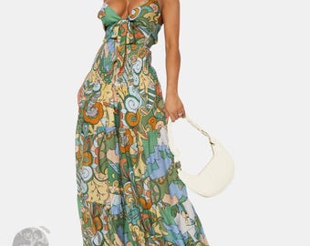 May include: A floor-length maxi dress featuring a multi-colored abstract print in shades of green, orange, yellow, and blue. The dress has a tie-front bodice, thin spaghetti straps, a smocked waist, and a tiered skirt. A white shoulder bag is included.