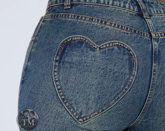 May include: High-waisted blue denim jeans featuring a prominent heart-shaped patch on the back. The medium-wash fabric displays a classic twill texture with visible stitching along the seams and the heart outline. A circular logo with the text TM is visible.