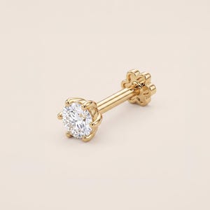 Könnte beinhalten: Ein goldener Ohrstecker mit einem einzelnen runden, im Brillantschliff geschliffenen, klaren Edelstein in einer sechs-Krappen-Fassung. Der gerade Stift endet in einem flachen, blumenförmigen Verschluss für einen sicheren Halt am Ohr.