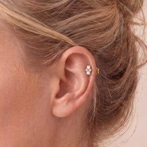 Può includere: Un orecchino da cartilagine color oro con tre piccole pietre trasparenti a taglio rotondo disposte in un gruppo verticale. L'orecchino è fissato nella parte superiore dell'orecchio e presenta una montatura metallica che sostiene le pietre.