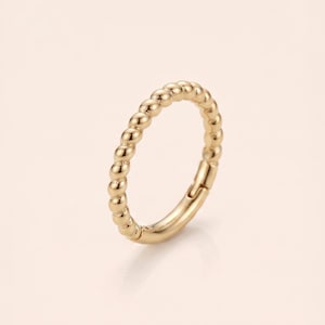 May include: A gold-toned ring featuring a continuous beaded or twisted texture along the outer band. The ring has a hinged mechanism visible on one side, allowing it to open and close securely. The metal surface has a polished, reflective finish.