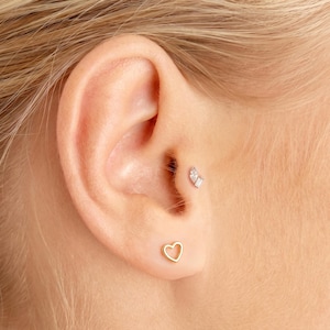 Könnte beinhalten: Ein goldener herzförmiger Ohrstecker befindet sich im Ohrläppchen, während ein kleiner silberfarbener Tragus-Ohrstecker mit zwei klaren, im Marquise- und Baguetteschliff geschliffenen Steinen im Tragus-Knorpel des Ohrs sitzt.