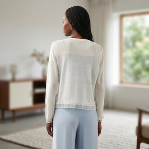 May include: A long-sleeved, off-white, semi-sheer knit cardigan featuring a round neckline and a delicate, pleated ruffle trim along the bottom hem. The garment is worn over a matching off-white camisole and paired with light blue trousers.