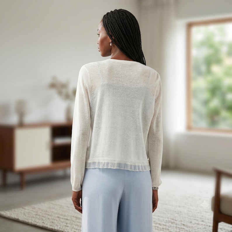 May include: A long-sleeved, off-white, semi-sheer knit cardigan featuring a round neckline and a delicate, pleated ruffle trim along the bottom hem. The garment is worn over a matching off-white camisole and paired with light blue trousers.