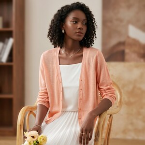 May include: A peach-colored long-sleeve cardigan featuring a V-neckline and a front button closure. The garment is worn over a white top and a white pleated skirt. The cardigan has ribbed cuffs and a lightweight knit texture throughout the fabric.
