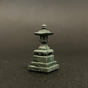 以下が含まれることがあります： 小さく、風化した緑色の日本の石灯籠。 ミニチュアの塔のような構造は、複数の層、ドーム型のトップ、正方形のベースが特徴です。 灯籠は暗い背景に配置され、その複雑なディテールを際立たせています。