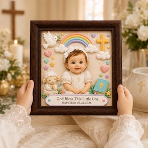 May include: A framed wall art piece featuring a central photograph of a baby in a white dress, surrounded by 3D-style decorations including a white dove, a rainbow, a yellow cross, a lamb, and a blue book. The text reads God Bless This Little One BAPTIZED 06.22.2026.