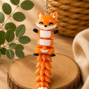 Könnte beinhalten: Ein orangefarbener Fuchsschlüsselanhänger mit einem silbernen Schlüsselring. Der Fuchs hat ein lächelndes Gesicht, schwarze Pfoten und eine weiße Brust. Der Schwanz ist ein stilisiertes Design. Der Schlüsselanhänger wird auf einer Holzscheibe präsentiert.