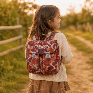 Può includere: Uno zaino con stampa floreale nei toni del rosso scuro, arancione e viola, dotato di due tasche frontali, fibbie in metallo argentato e il nome Charlotte ricamato in bianco sulla patta, indossato sopra un maglione in maglia color crema.