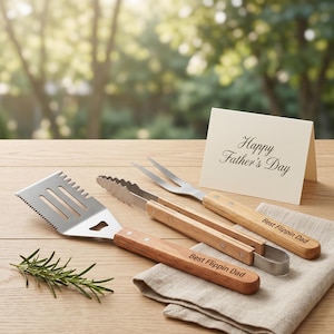 Puede incluir: Un juego de utensilios de parrilla de acero inoxidable con mangos de madera, que incluye una espátula, pinzas y tenedor, dispuestos sobre una superficie de madera. Se muestra una tarjeta con "Happy Father's Day". Las pinzas y la espátula están grabadas con "Best Flippin Dad".