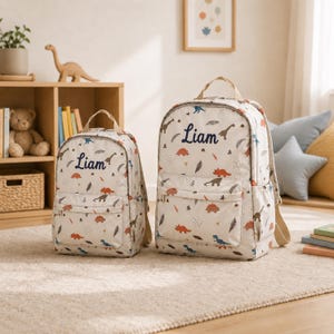 Peut inclure: Deux sacs à dos beiges avec un motif répétitif de dinosaures dans des tons orange, bleu et marron. Chaque sac possède une poche frontale zippée, une poignée supérieure et le prénom Liam brodé en bleu foncé sur le panneau avant.