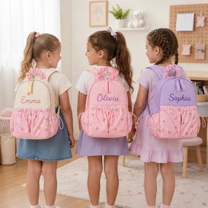 May include: Three backpacks featuring a top handle with a ruffled fabric detail, a main compartment with a zipper closure, and a front pocket with a pink pattern of rainbows and stars. Each bag is personalized with embroidered names in cursive script.