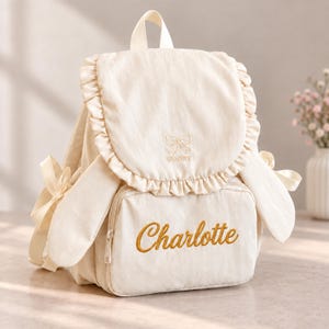 May include: A cream-colored backpack featuring a ruffled flap, long side panels resembling ears with tied ribbons, and a top handle. The flap displays an embroidered bow and the text SENIRY, while the front pocket features the name Charlotte.
